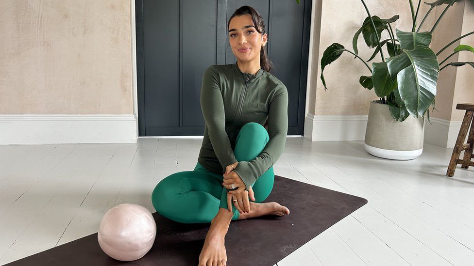Stef Williams is wearing teal gym leggings and a khaki longsleeve zipper. She is sitting on an exercise mat with a plant in the background.